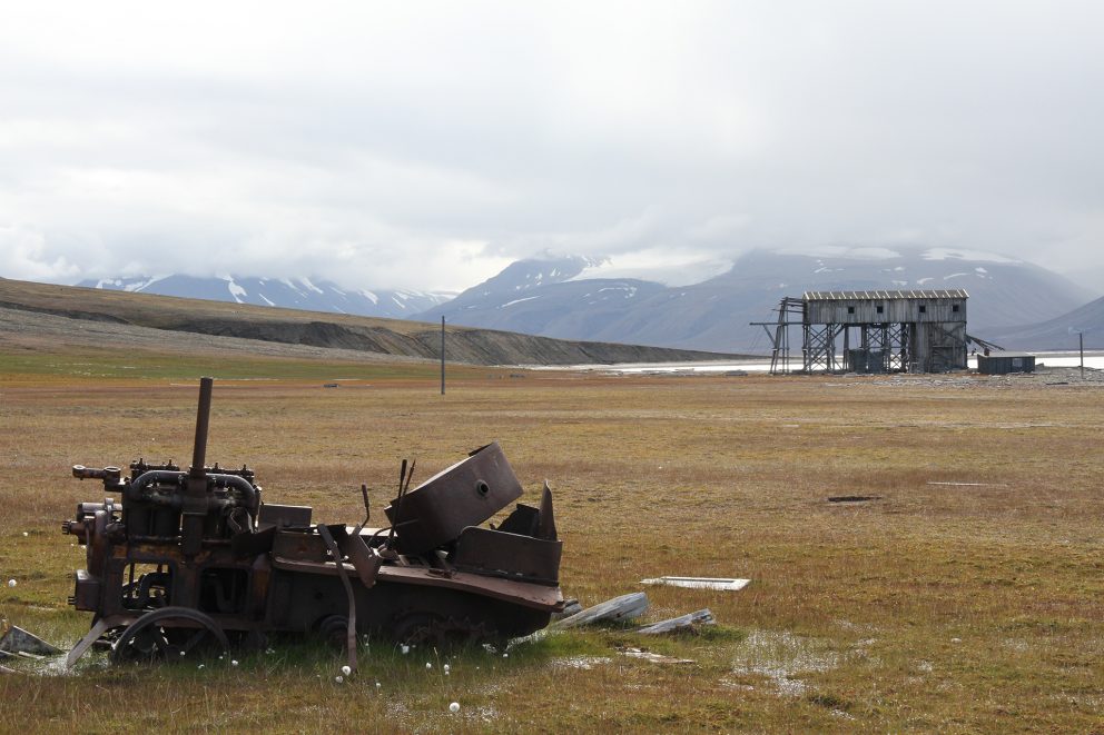 Goldt landskap med tundra og rustne rester etter en traktor i forgrunnen, taubanestasjonen med fjell, fjord og skyet himmel i bakgrunnen.