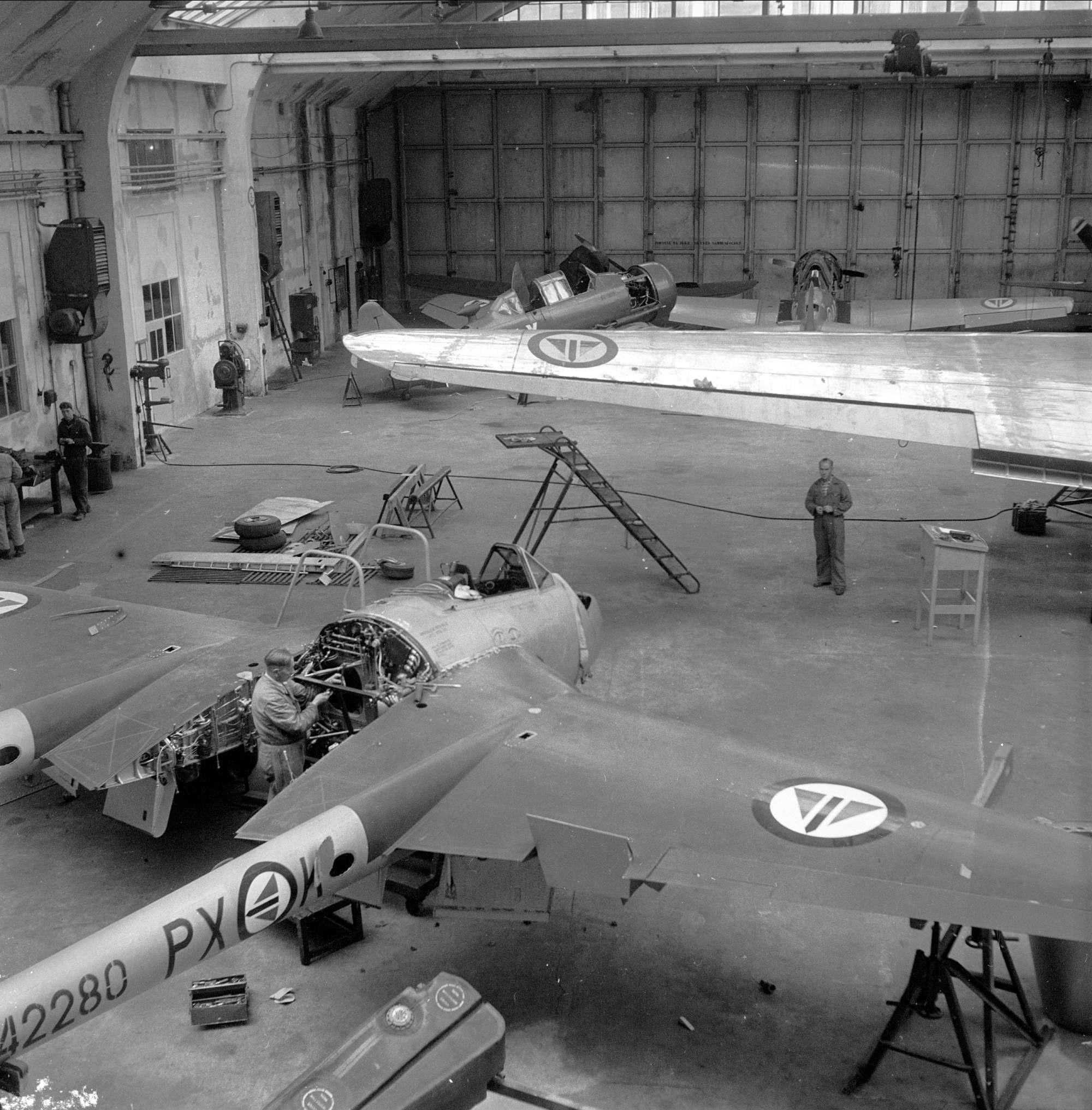 Bildet viser Vedlikehold av fly ved Kjeller, i forkant en DH Vampire, vingen til en DC3 Dakota og to skolefly av type North American T-6/J Harvard. Bildet er tatt i 1952 i forkant av 40-års markeringen til Kjeller. Foto er lånt fra Dagbladet/Norsk Folkemuseum