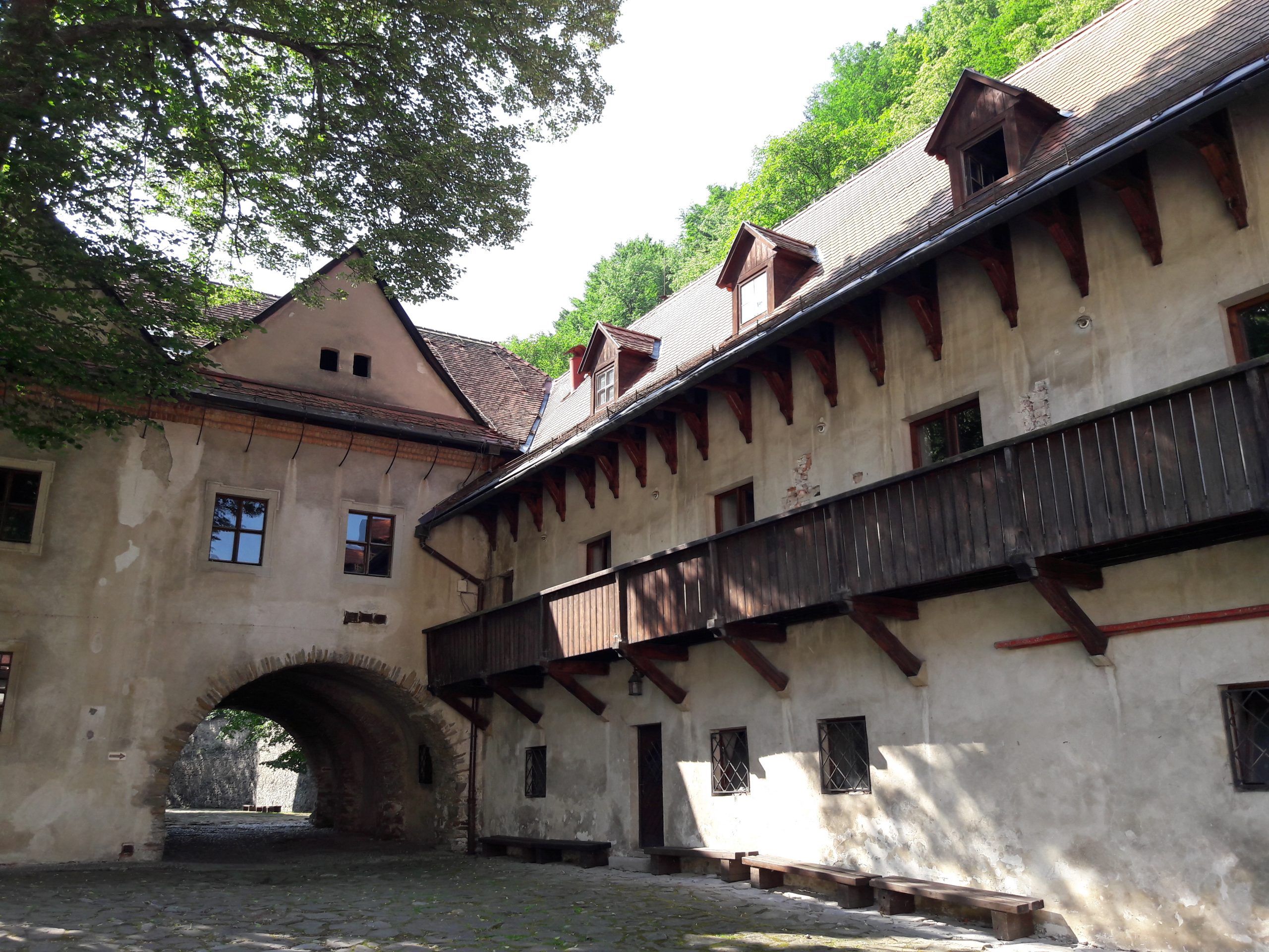 Historisk gårdsplass i stein med buet port, trebalkong og skråtak, omgitt av trær.