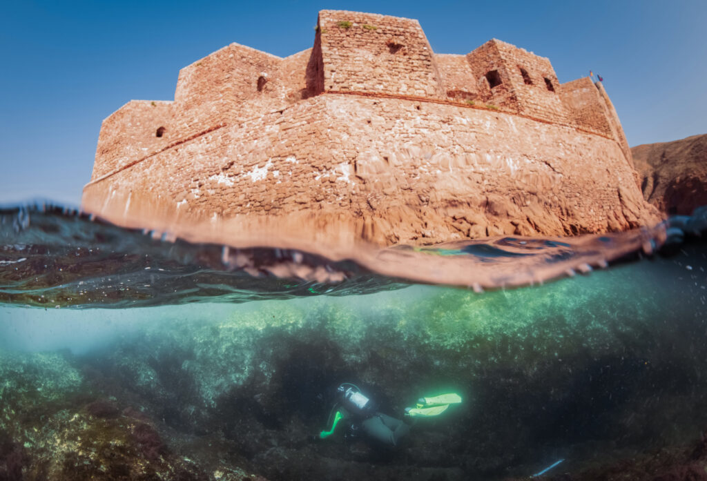 En dykker svømmer under vann i nærheten av en steinfestning, og bildet viser både det klare vannet under og den historiske strukturen over overflaten.