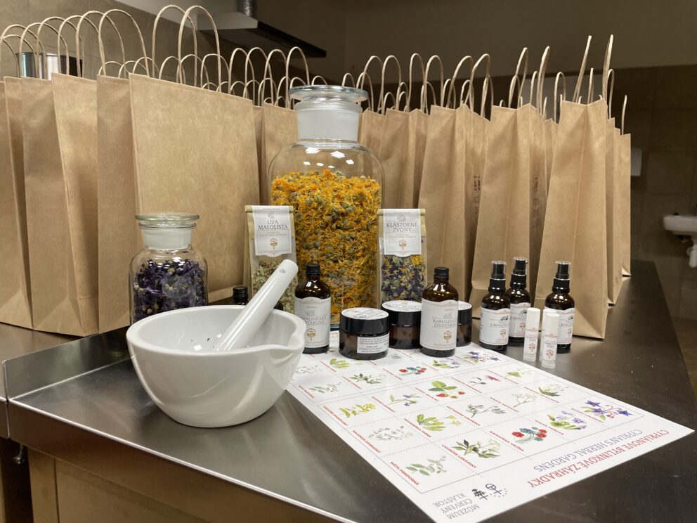 En utstilling med tørkede blomster, flasker, krukker og en morter på en benk, med papirposer og et botanisk kart i bakgrunnen.