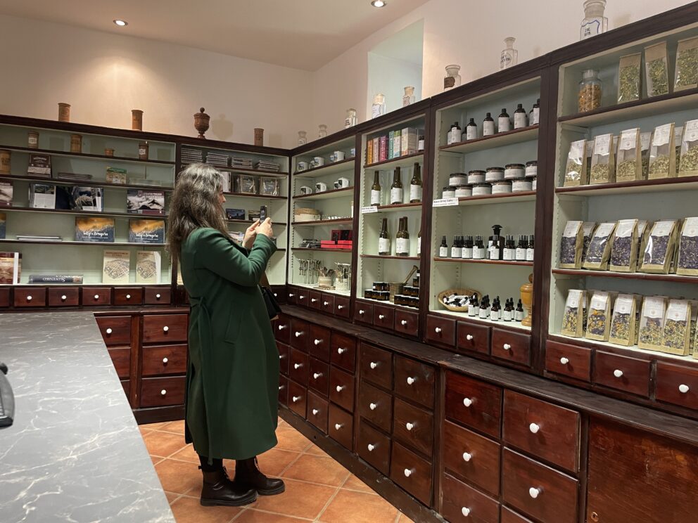 En kvinne i grønn frakk tar et bilde med mobilen i en apotekerbutikk med hyller fulle av krukker, flasker og pakker med urter.