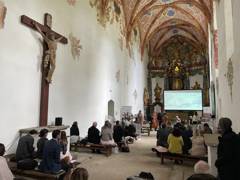 Folk sitter på benker inne i en utsmykket kirke, og deltar på et arrangement med et stort krusifiks, et utsmykket alter og en prosjektorskjerm som viser bilder foran.
