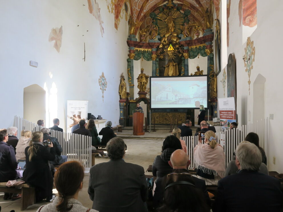 Folk sitter i en historisk kirke og overværer en presentasjon med en skjerm som viser et bilde av bygningen. Foran er det utsmykkede alteret og de religiøse statuene synlige.