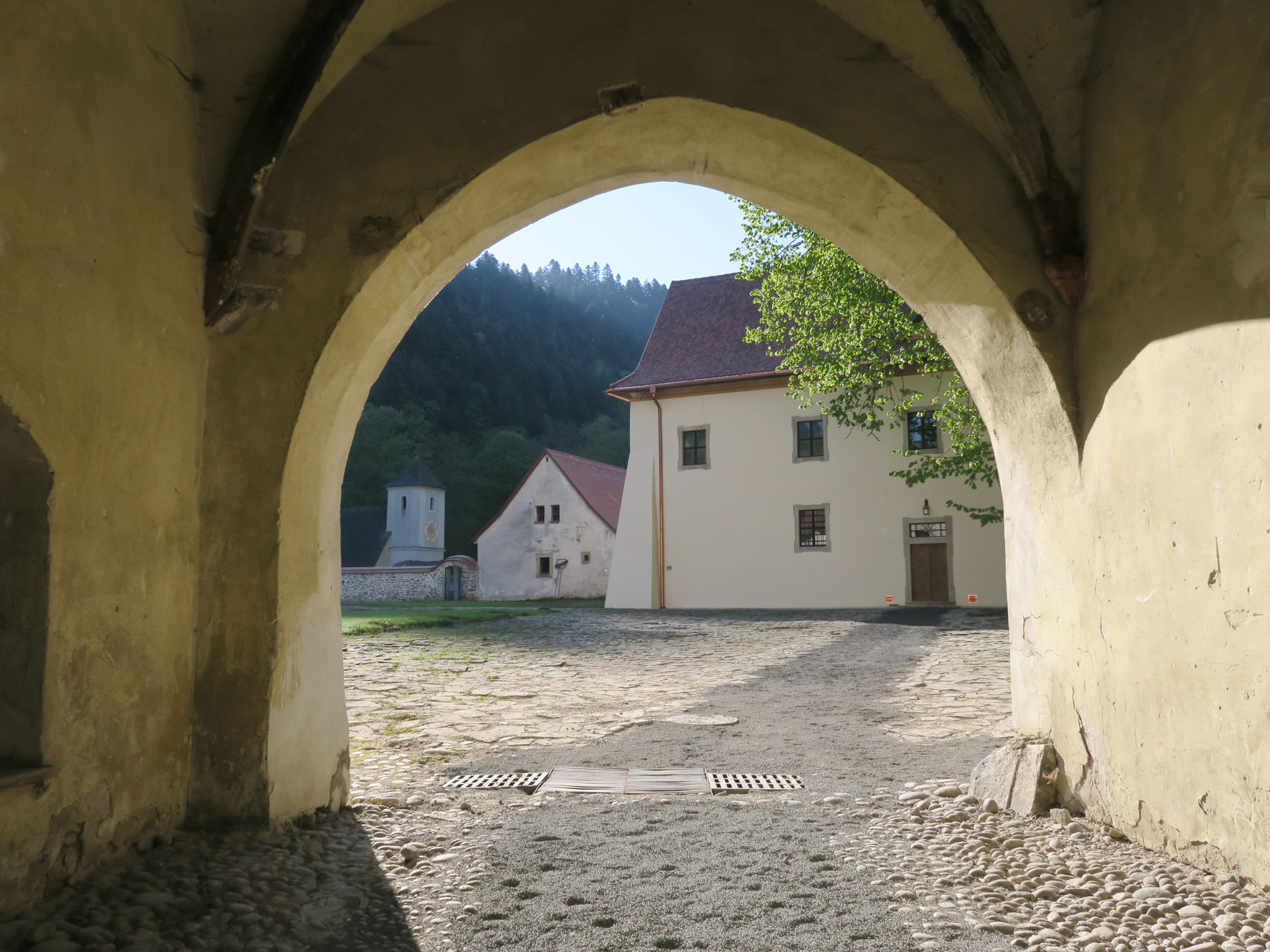 Utsikt gjennom en steinbue til en solbelyst gårdsplass med brostein, historiske bygninger og grønne åser i bakgrunnen.
