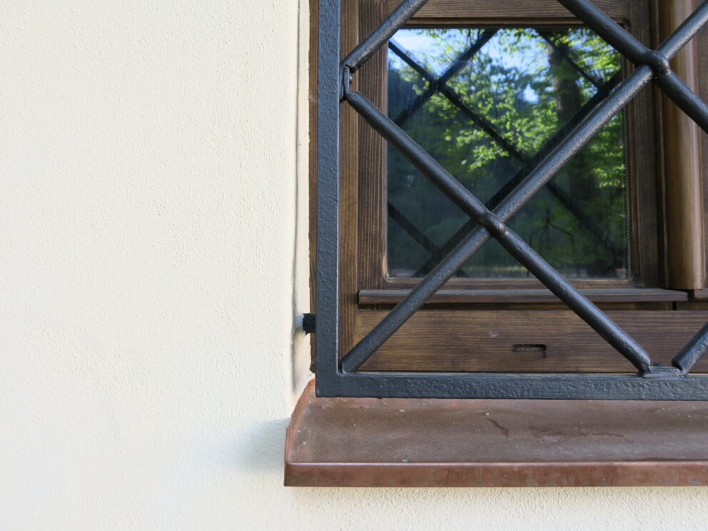 Nærbilde av et vindu med treramme og sorte sikkerhetsgitter i metall, innfelt i en lys vegg, som reflekterer grøntområdene utenfor.