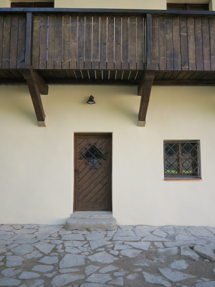 En tredør med diagonalt mønster og et lite vindu ved siden av på en kremfarget vegg, toppet av en trebalkong.