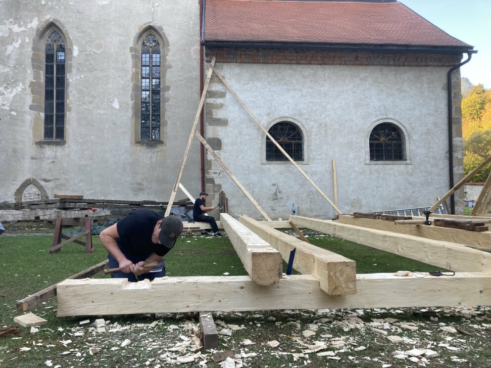 To personer arbeider på en bindingsverkskonstruksjon utendørs ved siden av en gammel steinbygning med høye, buede vinduer. På gresset ligger det trespon spredt utover.