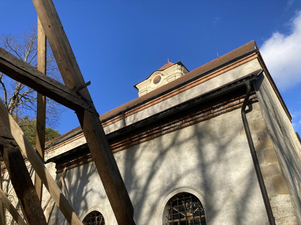 En steinbygning med buede vinduer og rødt tak i sollys, delvis innrammet av trebjelker, mot en klarblå himmel.