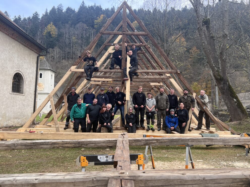 En gruppe mennesker poserer i og rundt en delvis oppbygd A-ramme av tre utendørs, med trær og en bygning i bakgrunnen.