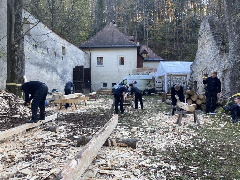 Flere personer arbeider med tre utendørs, med håndverktøy og benker, omgitt av spon, tømmerstokker og historiske steinbygninger i et skogsområde.