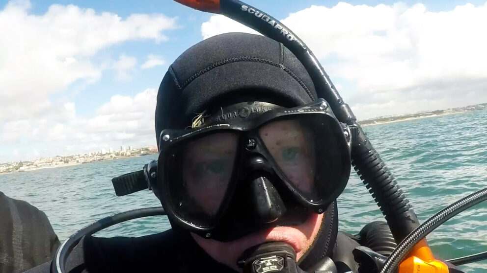 Person i fullt dykkerutstyr med maske og snorkel som tar en nærbilde-selfie på vannet, med kystlinje og skyer i bakgrunnen.