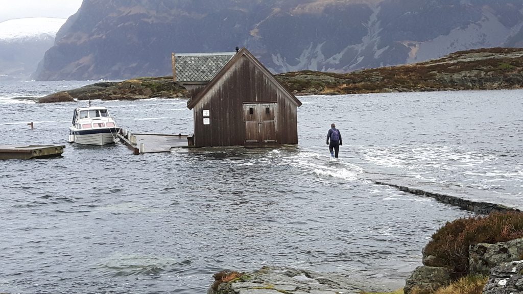 Beredskapsplanar: En person går på en delvis nedsenket sti mot et lite båthus i tre omgitt av vann, med en båt fortøyd i nærheten og fjell i bakgrunnen.