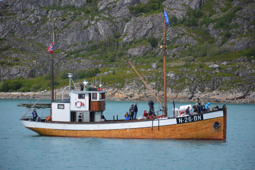 Fiskefartøy med mannskap om bord liggende på stille vann med dramatisk fjellside i bakgrunnen