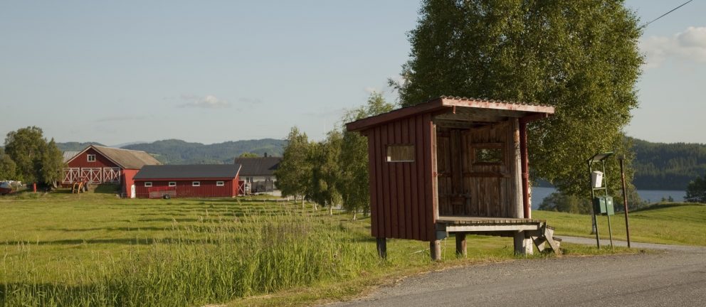 Hvorfor freder vi kulturmiljø og kulturminner? 2 En liten bussholdeplass i tre står ved en landevei, med røde låver, gressplener og trær i bakgrunnen under en klar himmel.
