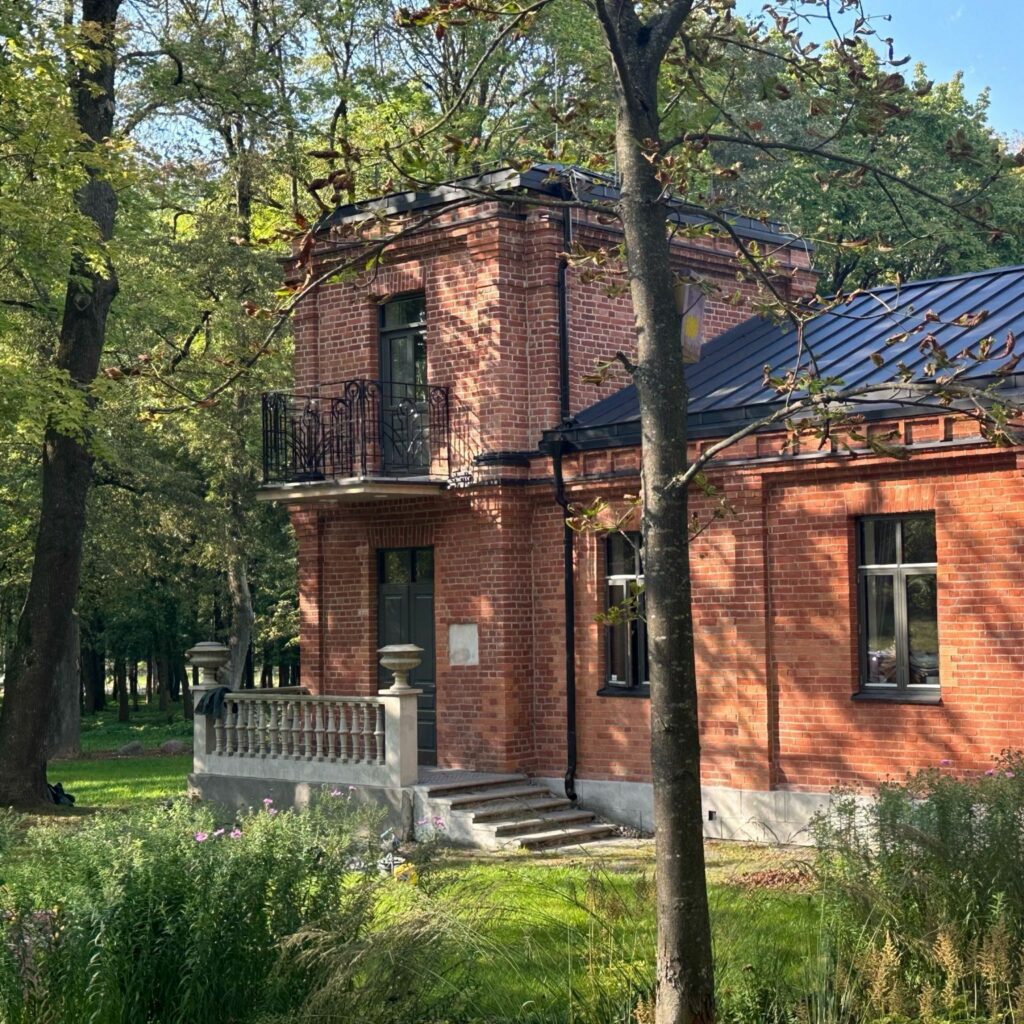 En toetasjes rød mursteinsbygning med store vinduer og balkong, omgitt av trær og grøntområder på en solrik dag.