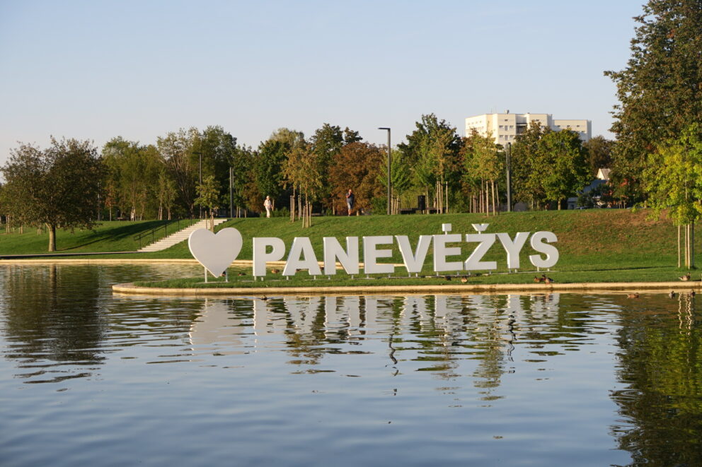 Det store hvite I ♥ Panevėžys-skiltet står ved en rolig vannkant, med trær og moderne bygninger i bakgrunnen under en klar himmel.