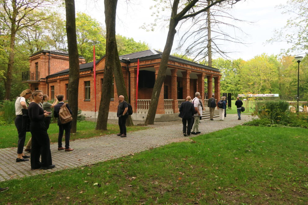 Folk står og snakker på en asfaltert sti i nærheten av en rød mursteinsbygning omgitt av trær og grøntområder i et parkanlegg.