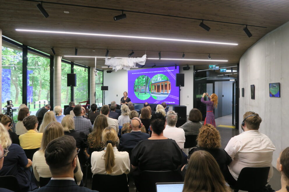 En gruppe mennesker sitter i et moderne konferanserom og ser på en foredragsholder som presenterer lysbilder om en bygning, mens en annen person fotograferer begivenheten.