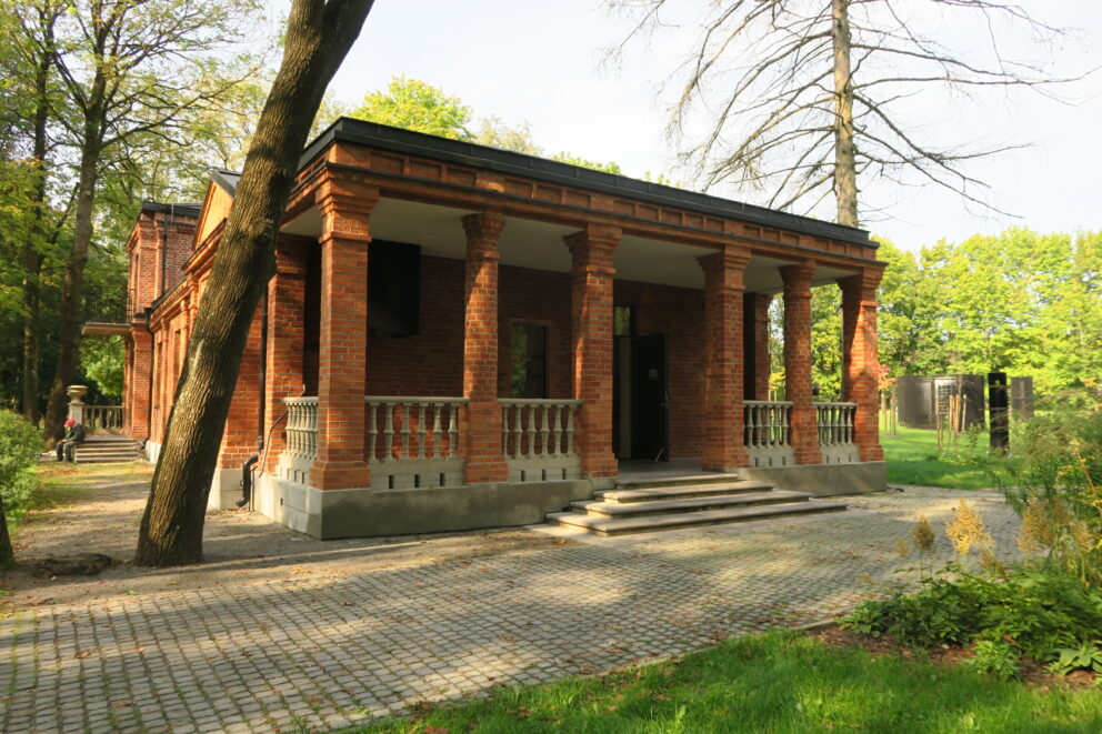 En rød mursteinsbygning med søyler og en liten trapp, omgitt av trær og grøntområder på en solrik dag.