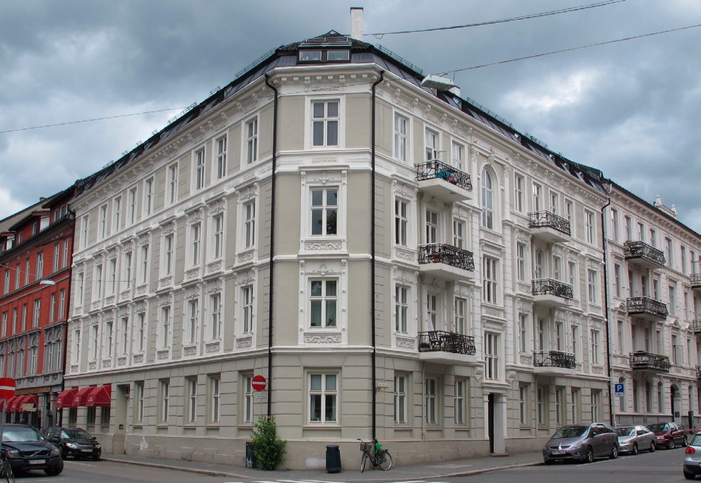 En fireetasjes hjørneboligblokk med utsmykkede hvite detaljer, balkonger og flere vinduer, omgitt av parkerte biler og en overskyet himmel over seg.