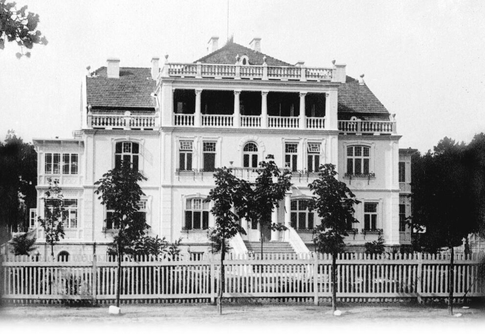 Sort/hvit fotografi av en storslagen villa i flere etrasjer, med overbygd balkong og doble vinduer. Hvitt stakittgjerde og trær i forgrunnen.