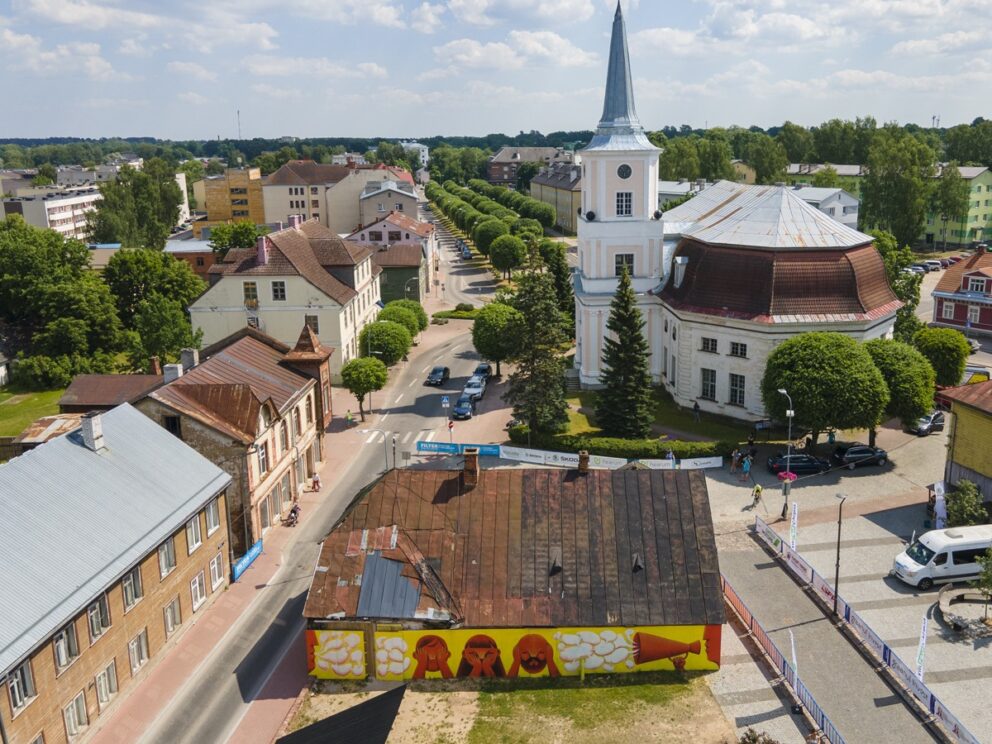 Oversiktsbilde av en bykjerne med kirke, gamle bygninger, veier, trør og en nedslitt trebygning i midten.