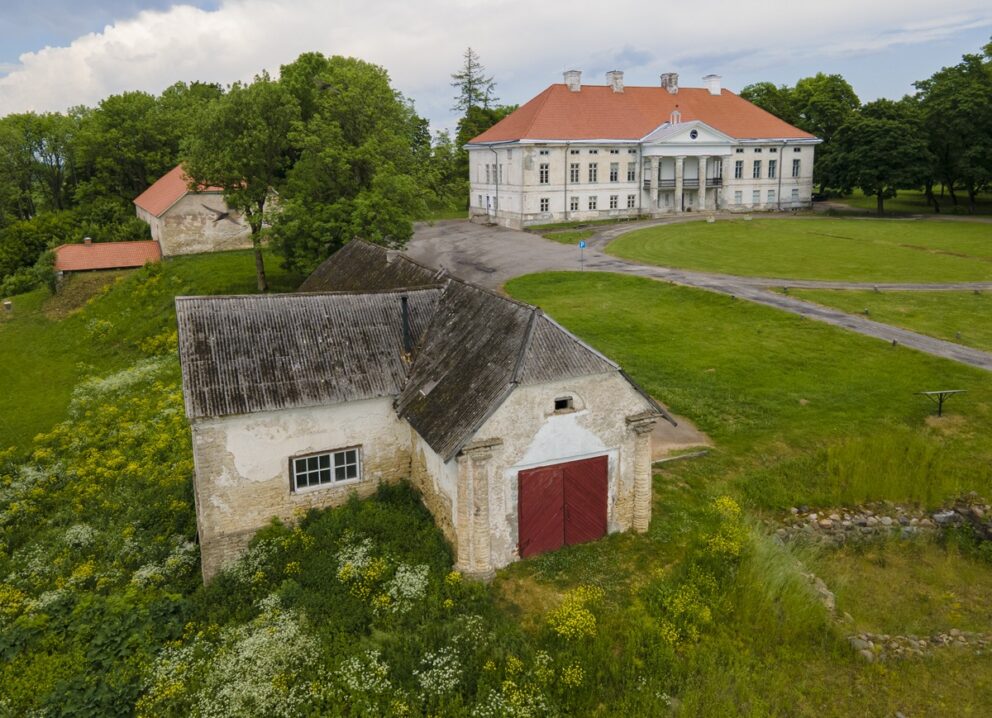 En gammel murbygning med røde dører, liggende i forgrunnen av en noe forfallen herregårdsbygning, med grønne plener, hvite og gule markblomster og trær.