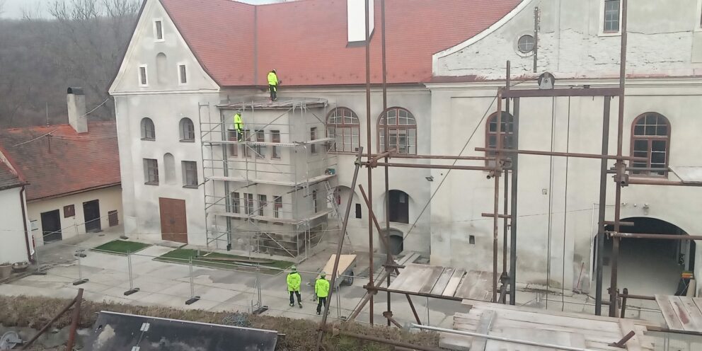 Restaureringsarbeid i gang på en herregård, med stilaser, arbeidere som jobber på tak og vegger.