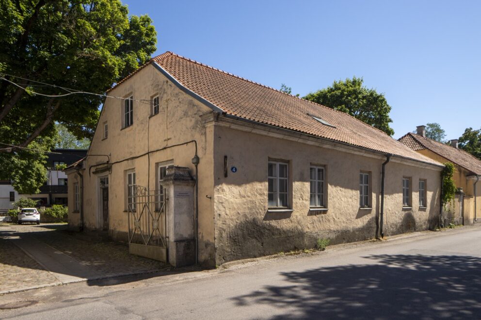 En slitt, gul murbugning beliggende i en gate med gamle hus, med brostein foran og åpen smijernsport.