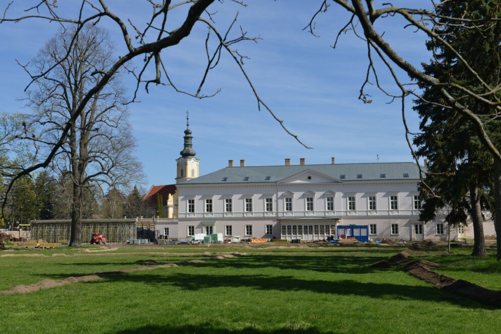 En hvit herregårdsbygniing med en gul kirke i bakgrunnen, med plen og høye trær.