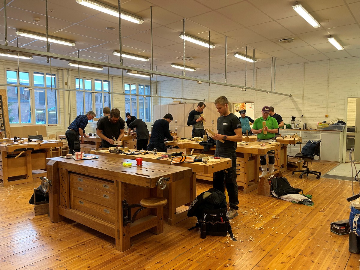 Flere personer jobber med trebearbeidingsprosjekter ved individuelle benker i et godt opplyst verksted med tregulv og store vinduer.