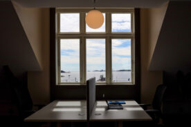 To tomme kontorpulter med stoler vender mot et stort vindu med utsikt over havet og himmelen, med en bærbar PC og en notatbok på det ene skrivebordet og en rund taklampe over.