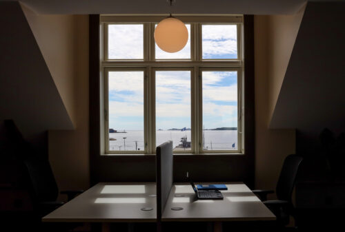To tomme kontorpulter med stoler vender mot et stort vindu med utsikt over havet og himmelen, med en bærbar PC og en notatbok på det ene skrivebordet og en rund taklampe over.