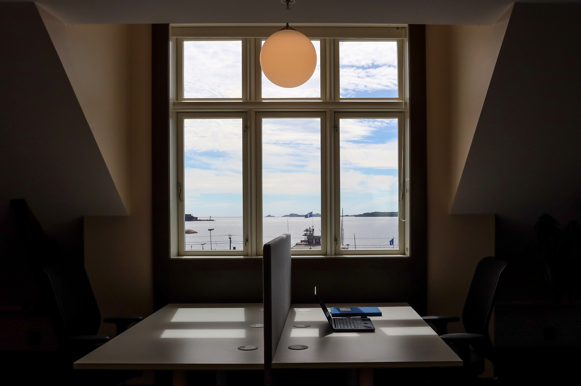 To tomme kontorpulter med stoler vender mot et stort vindu med utsikt over havet og himmelen, med en bærbar PC og en notatbok på det ene skrivebordet og en rund taklampe over.