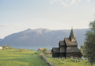 En trekirke med spiss tak står på en gresskledd kirkegård, med utsikt over en fjord og store åser i bakgrunnen under en klar himmel.