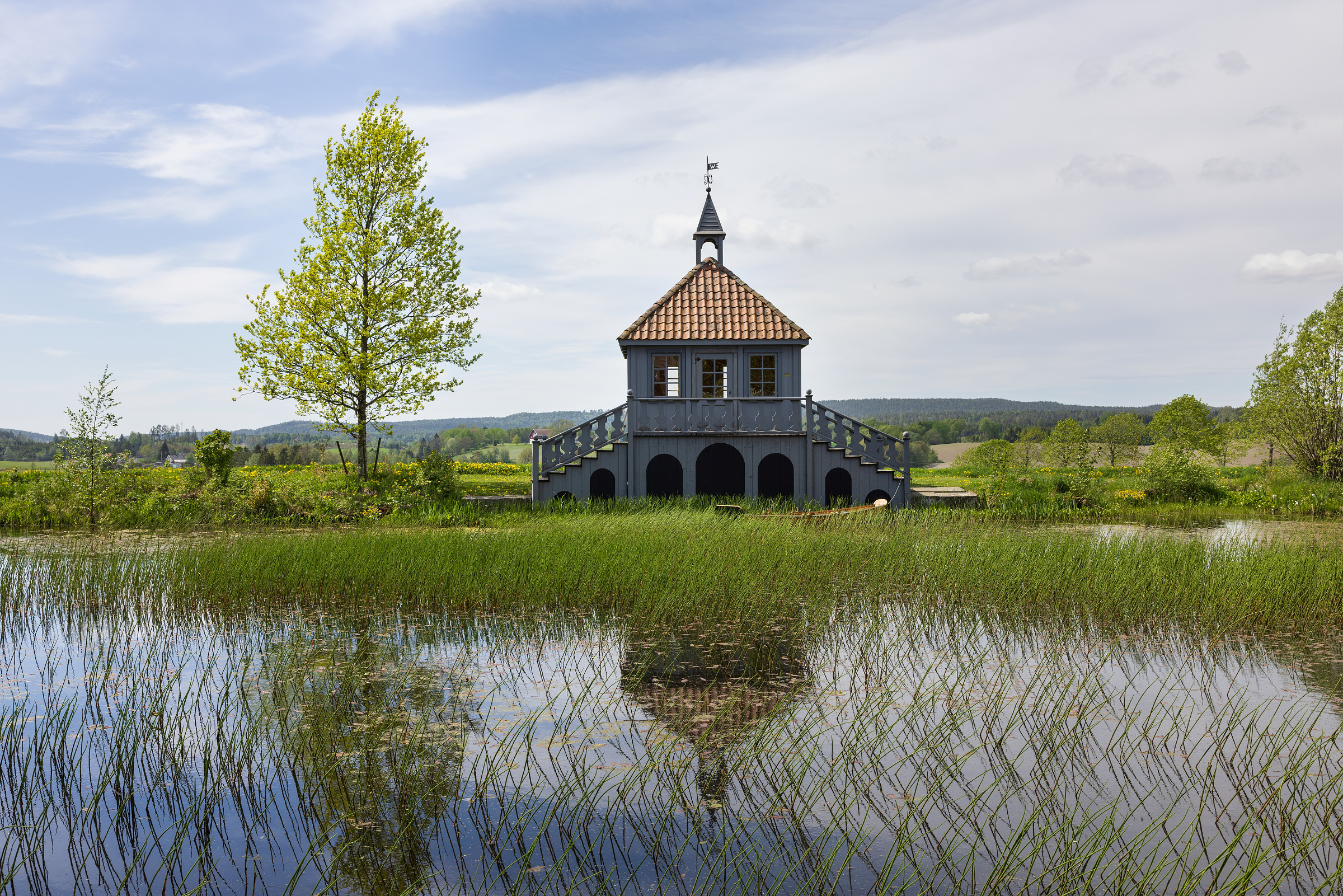 Det historiske lysthuset med dammen med siv på Spydeberg prestegård i Indre Østfold, i sommerhalvåret med grønt gress og blader på treet.
