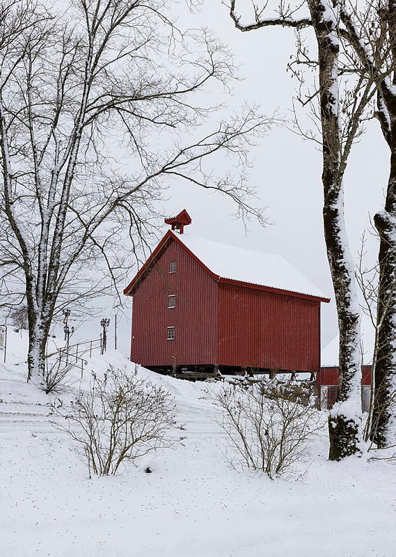 Rødt stabbur, hvit snø og trestammer på Spydeberg prestegård