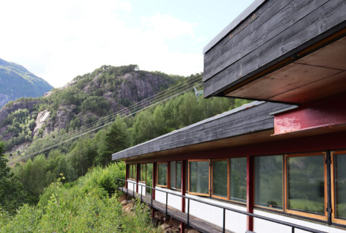 En moderne bygning med store vinduer og et eksteriør i tre står foran skogkledde fjell og kraftledninger på en solrik dag.
