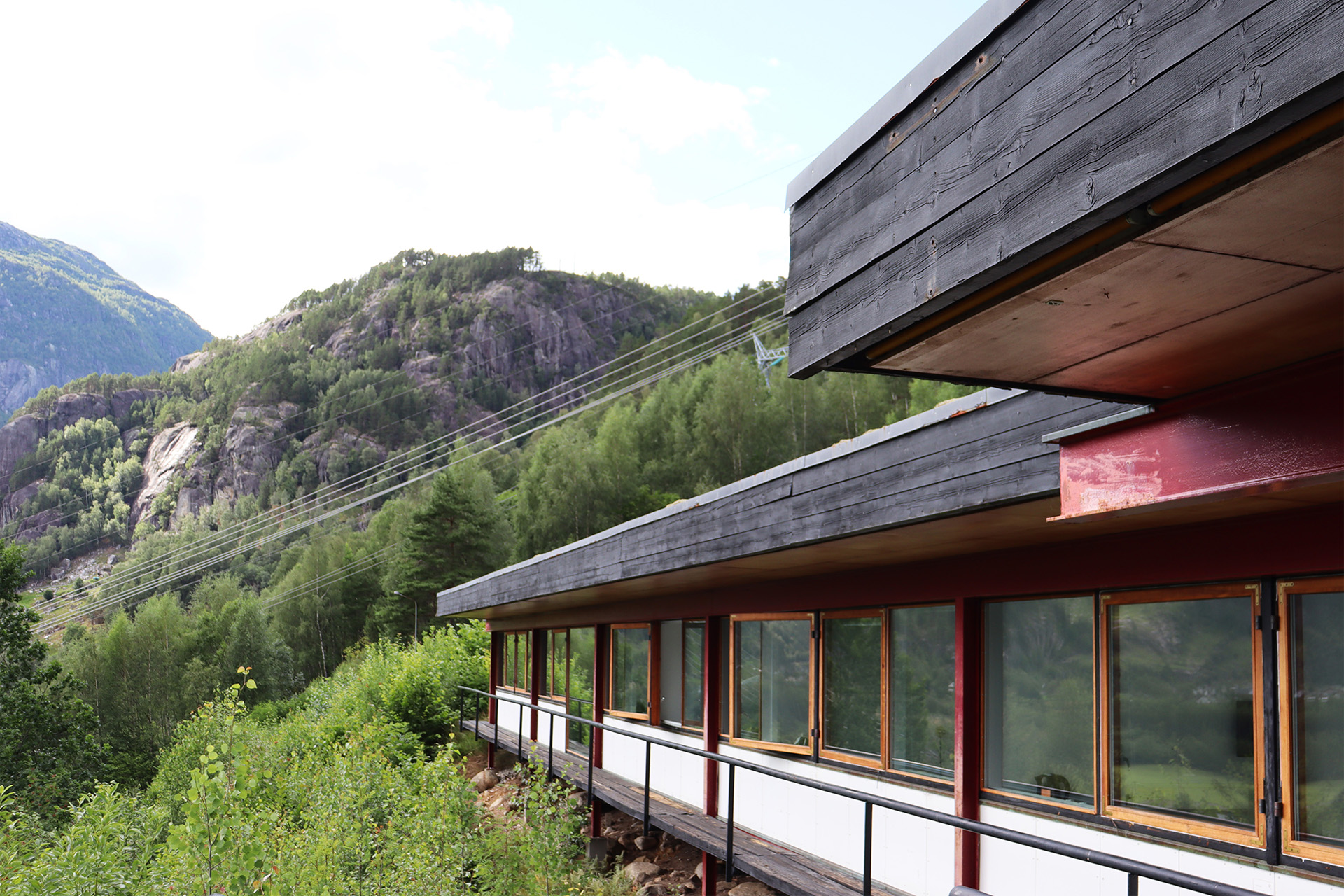 En moderne bygning med store vinduer og et eksteriør i tre står foran skogkledde fjell og kraftledninger på en solrik dag.