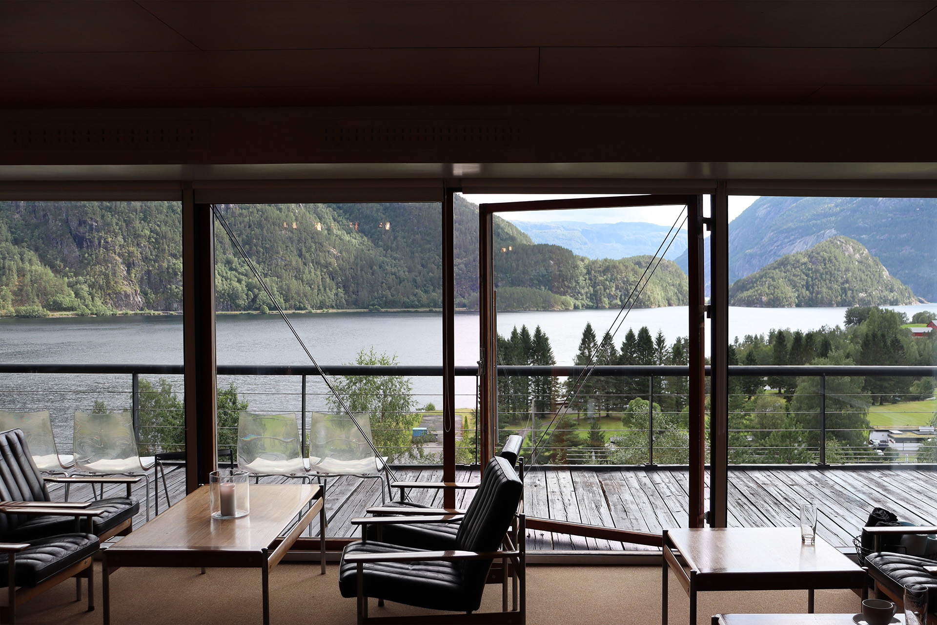 En moderne salong med svarte stoler og bord har utsikt over en innsjø og fjell gjennom store glassvinduer og en åpen dør som fører til en trebalkong.
