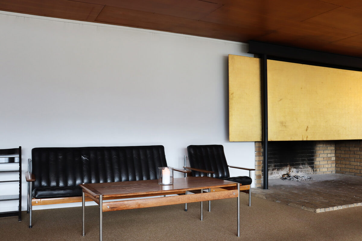 En minimalistisk stue med svart skinnsofa, lenestol, salongbord i tre og peis med et stort gult panel over.