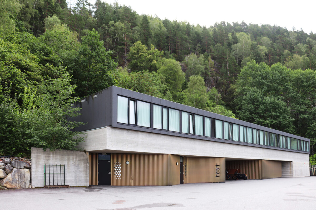 En moderne, rektangulær to-etasjers bygning med store vinduer og en betongbase er satt mot et bakteppe av tette grønne trær og skogkledd åsside.