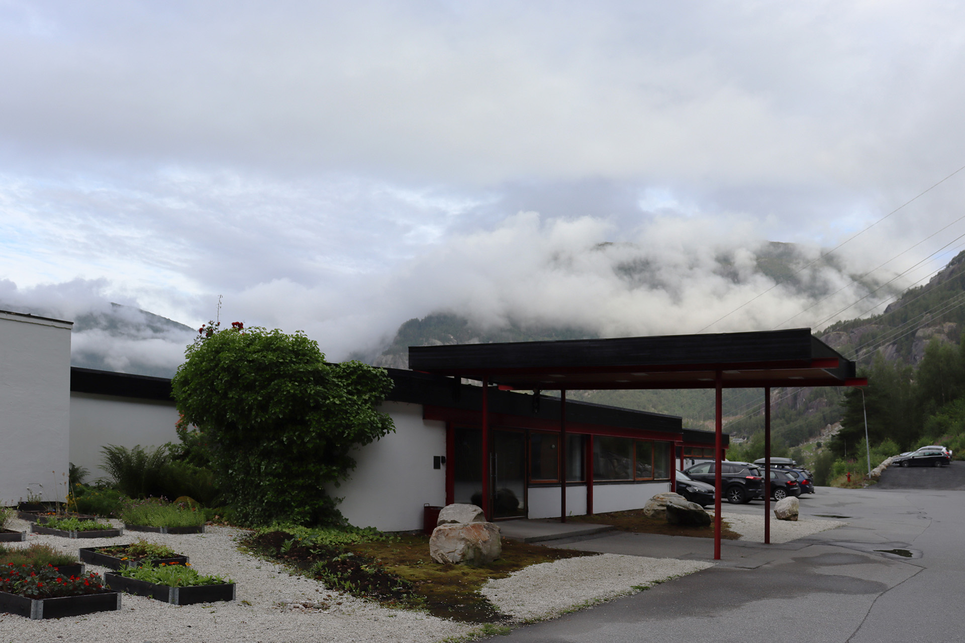En moderne, lav bygning med store vinduer og flatt tak står foran tåkete, skydekte fjell. Flere parkerte biler er synlige nær inngangen.