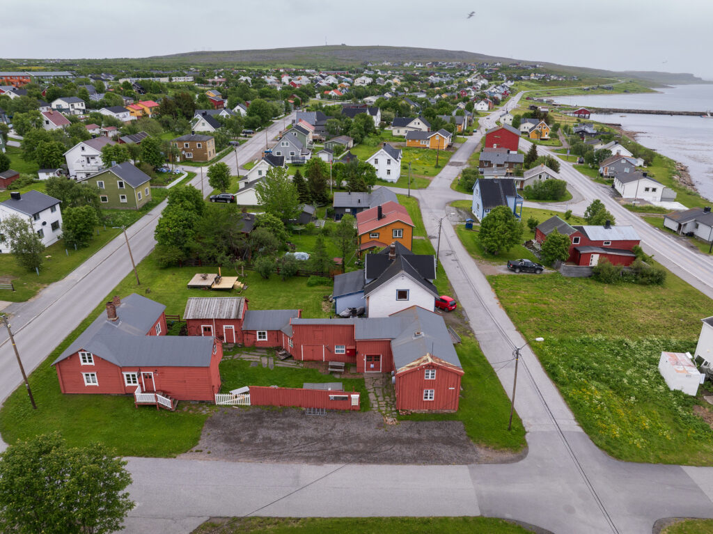 Luftfoto av Vadsø med fargerike hus, asfalterte veier, grønne plener og en strandlinje synlig på høyre side under en overskyet himmel.