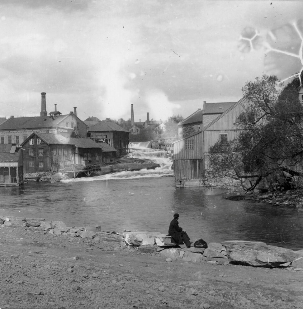 En person sitter på steiner ved en elv med flere industribygg og en foss i bakgrunnen.