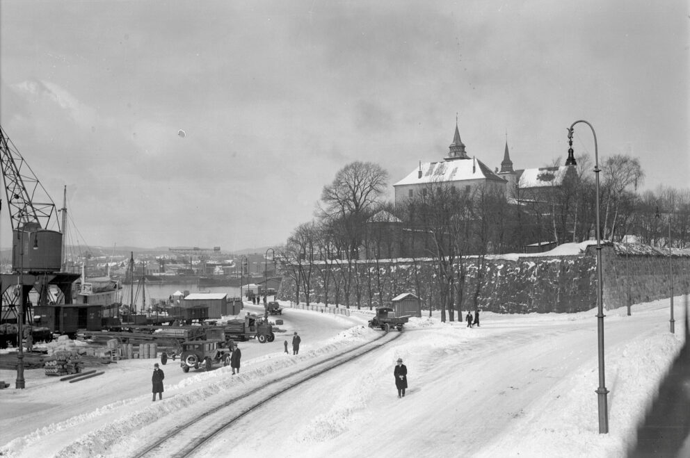 Snødekt gatebilde med folk som går og kjøretøy på en vei ved siden av en historisk bygning. Togspor synlige, og en industrikran i bakgrunnen. Trær og overskyet himmel fullfører scenen.