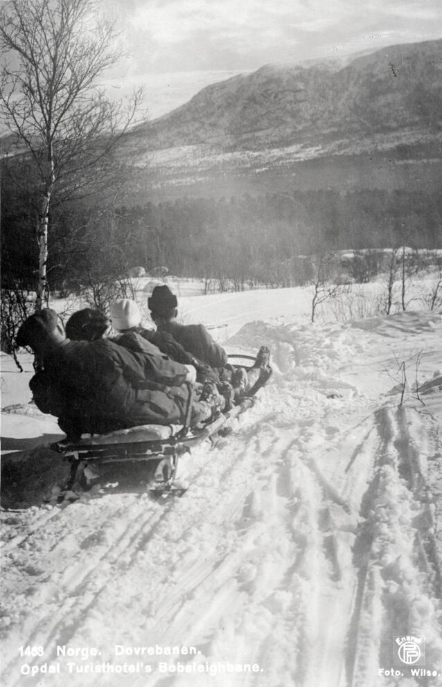 Fire personer kjører bobslede på en snødekt sti omgitt av trær og fjell, med spor i forgrunnen.