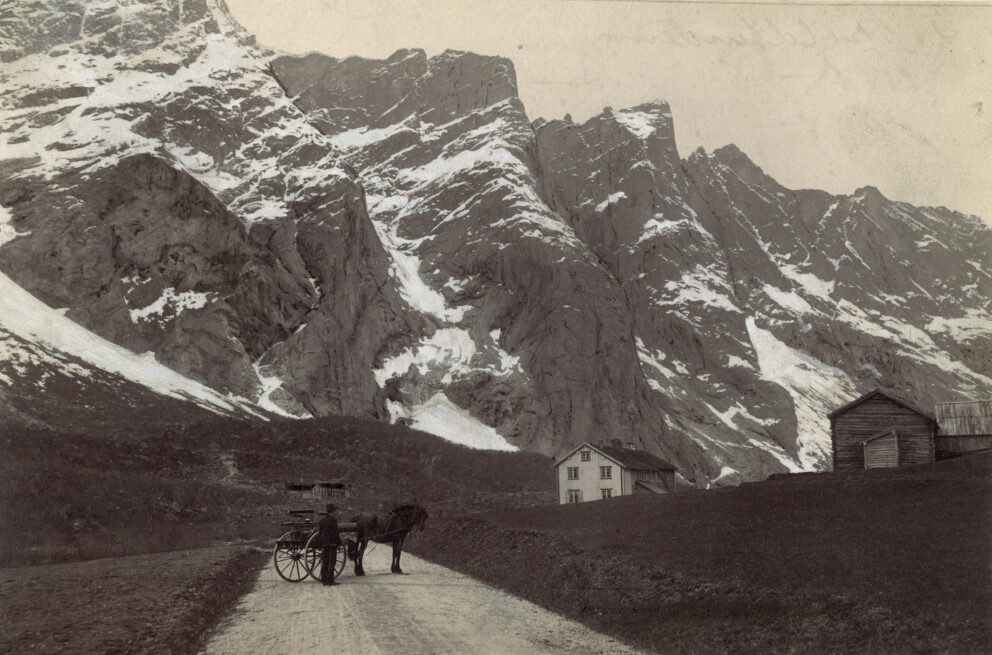 Hestevogn med to personer på fjellvei, med snødekte topper og trehusbebyggelse i bakgrunnen.