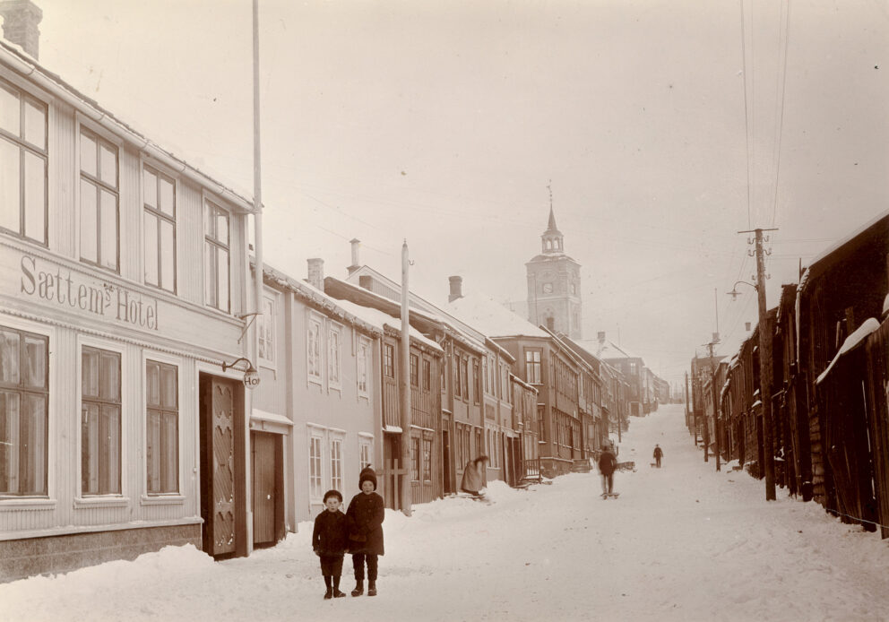 Et snørikt gatebilde med to barn i forgrunnen. Bygninger langs veien, og en kirke med et klokketårn er synlig i det fjerne.